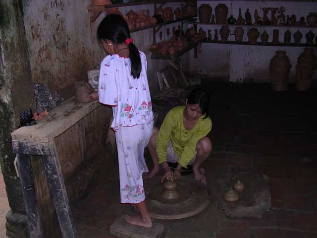 The girl standing is operating the wheel with her foot while decorating her partner's work